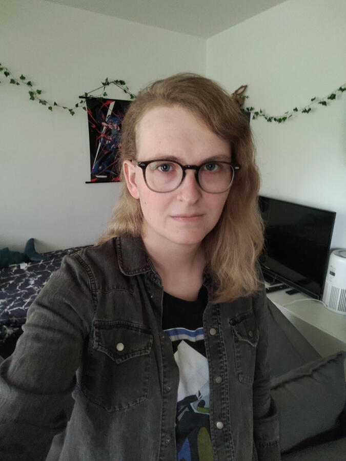 Jade looks into the camera with a neutral or concerned expression. She is wearing a shirt with some print and a gray denim jacket. In the background you can see a bed, a TV, a manga poster and fairy lights on the wall.