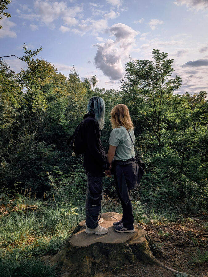 Mia and Jade looking at the clouds Mia and Jade are standing on a tree stump in a forest, holding hands and looking at the clouds in the sky. Mia's hair is dyed blue, Jade's is naturally blond. They are wearing casual summer clothes.