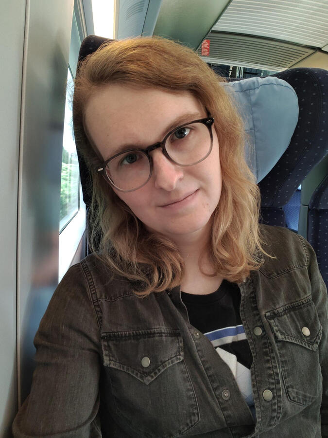 Another selfie of Jade in the seat of a train. She looks into a camera with a neutral expression. She is wearing the same clothes as in the first photo: a shirt with some logo and a gray denim jacket.